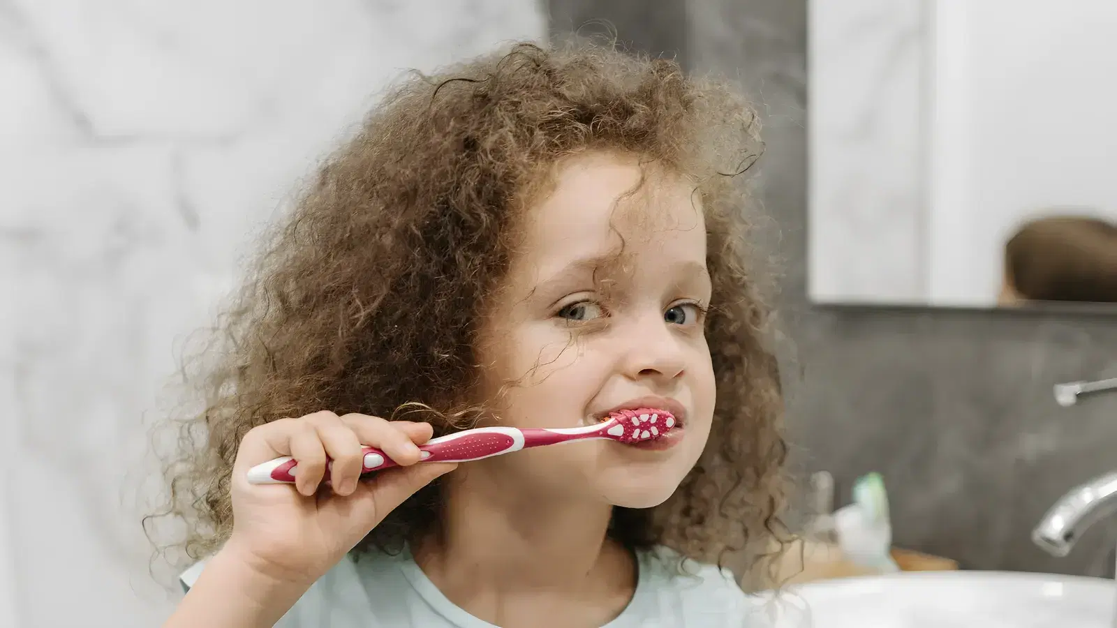 A young girl brushing her baby teeth with a pink toothbrush at the bathroom basin