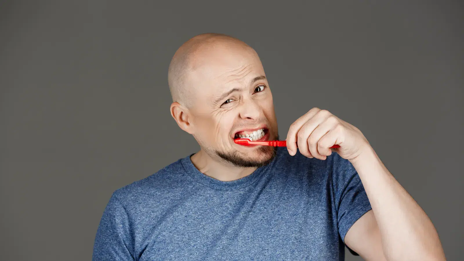 A man wincing as he brushes his teeth with a red toothbrush, jaw tensed against a sore gumline