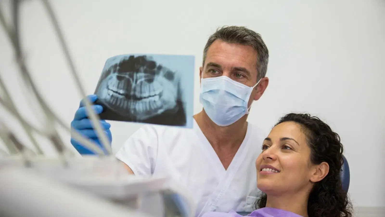 A dentist in a mask holding up a full-mouth X-ray for a smiling patient before a wisdom tooth removal